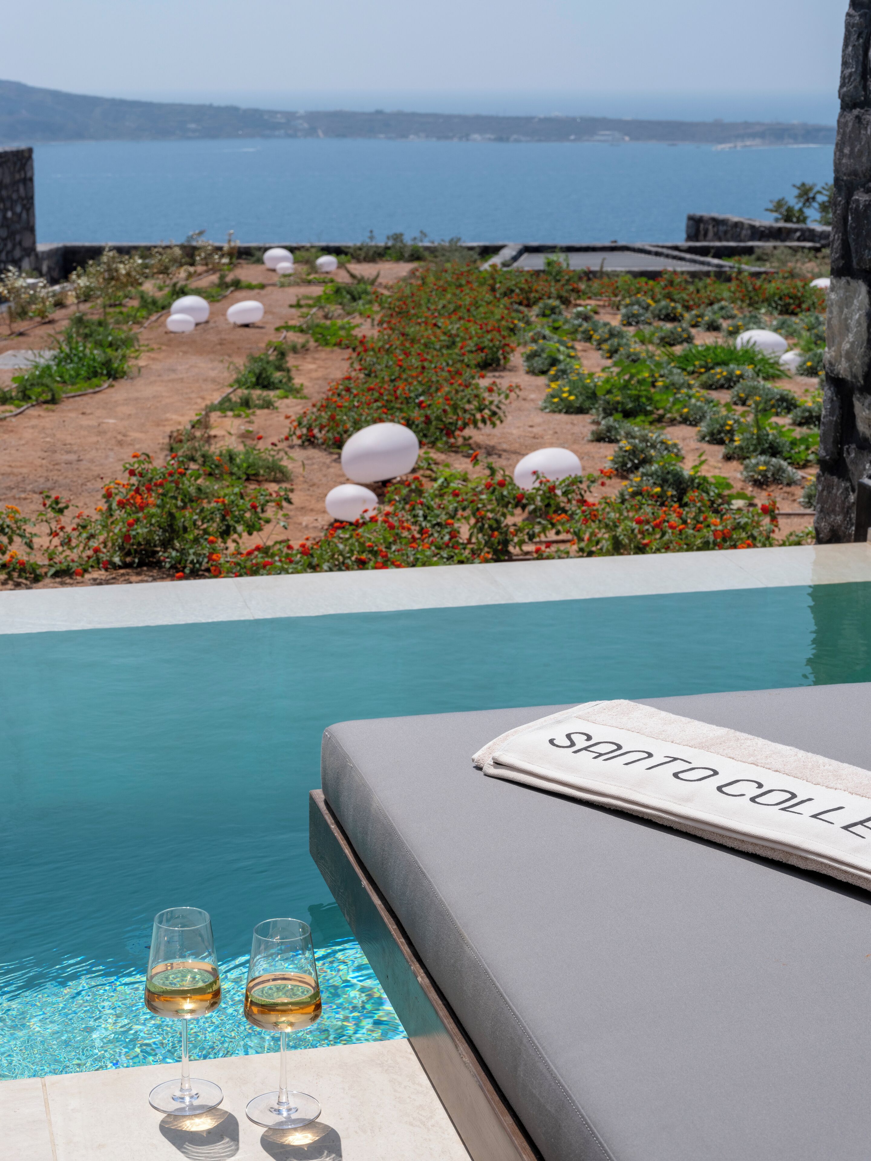 A poolside area with a lounge chair and two glasses of white wine, a sign reading "Santo Colle", and a view of a landscaped garden with white spherical decorations and red flowers.
