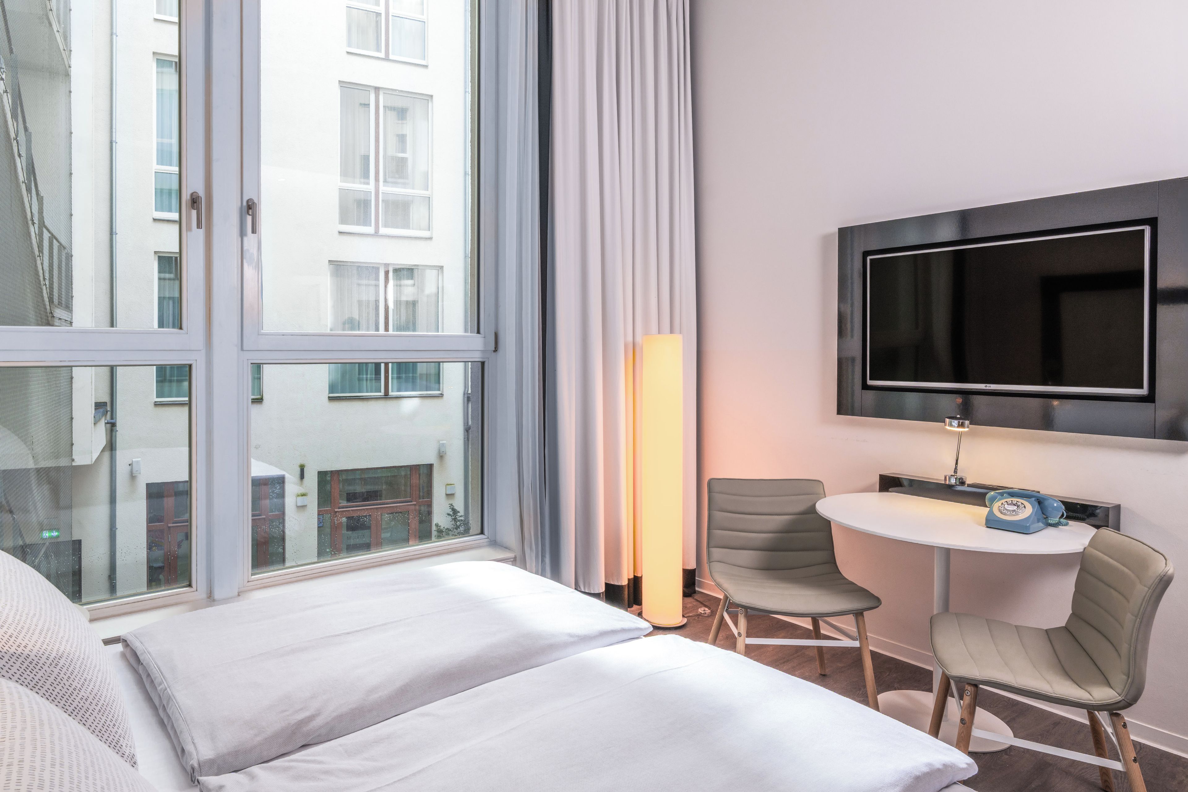 A hotel room with a bed, a TV mounted on the wall, a small table with chairs, and a window with curtains.