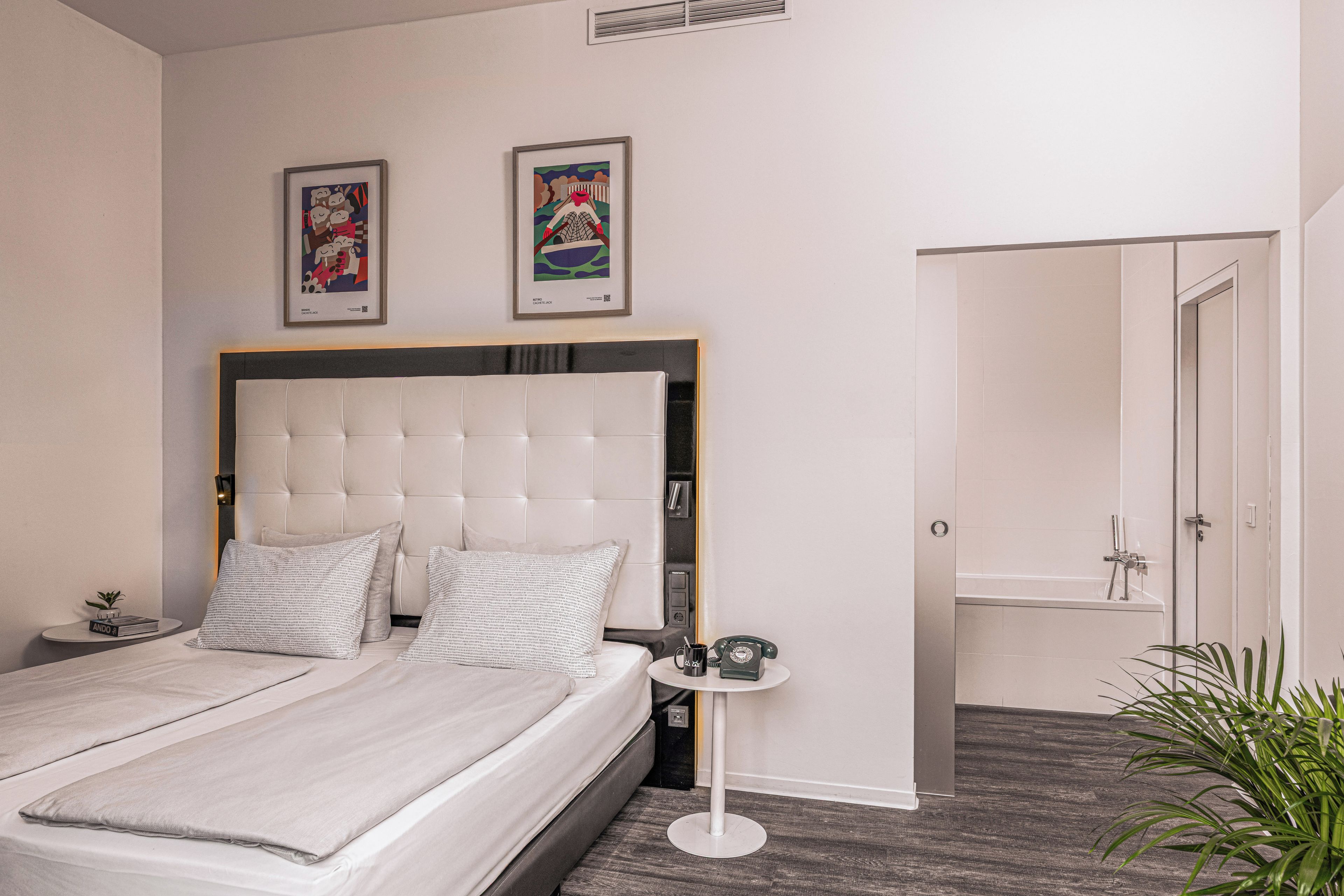 A modern bedroom with a large bed, bedside tables, a small side table, a framed picture on the wall, and a door to the bathroom.
