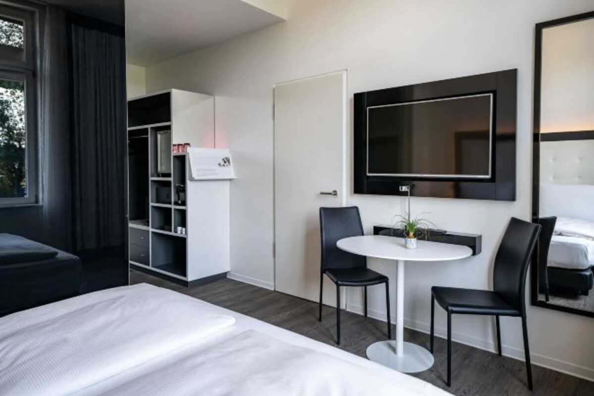 Ett modernt hotellrum med en säng, en tv som är monterad på väggen, ett litet runt bord med två stolar och en garderob.