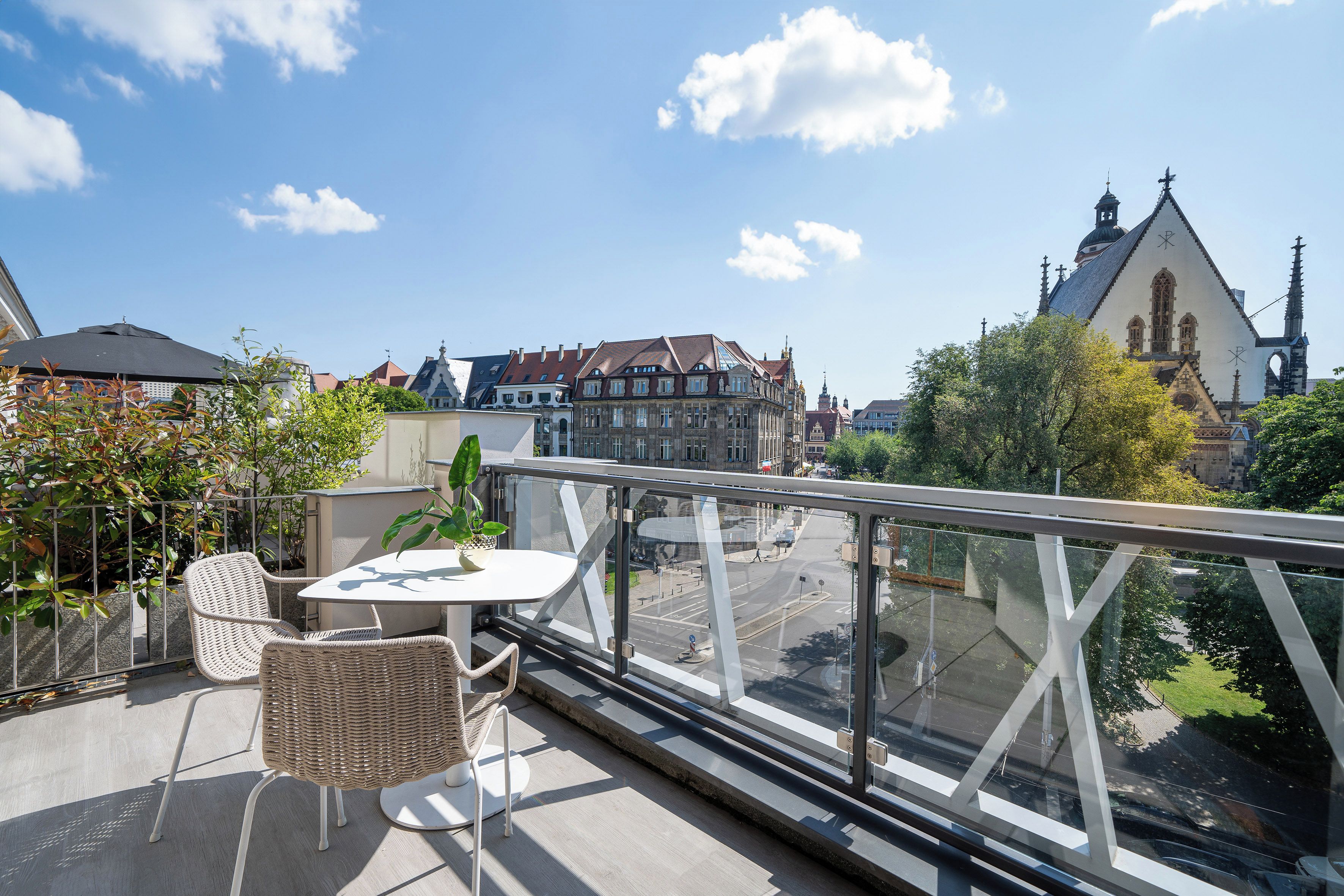 A balcony with wicker chairs and a table, overlooking a cityscape with historic buildings and a church.