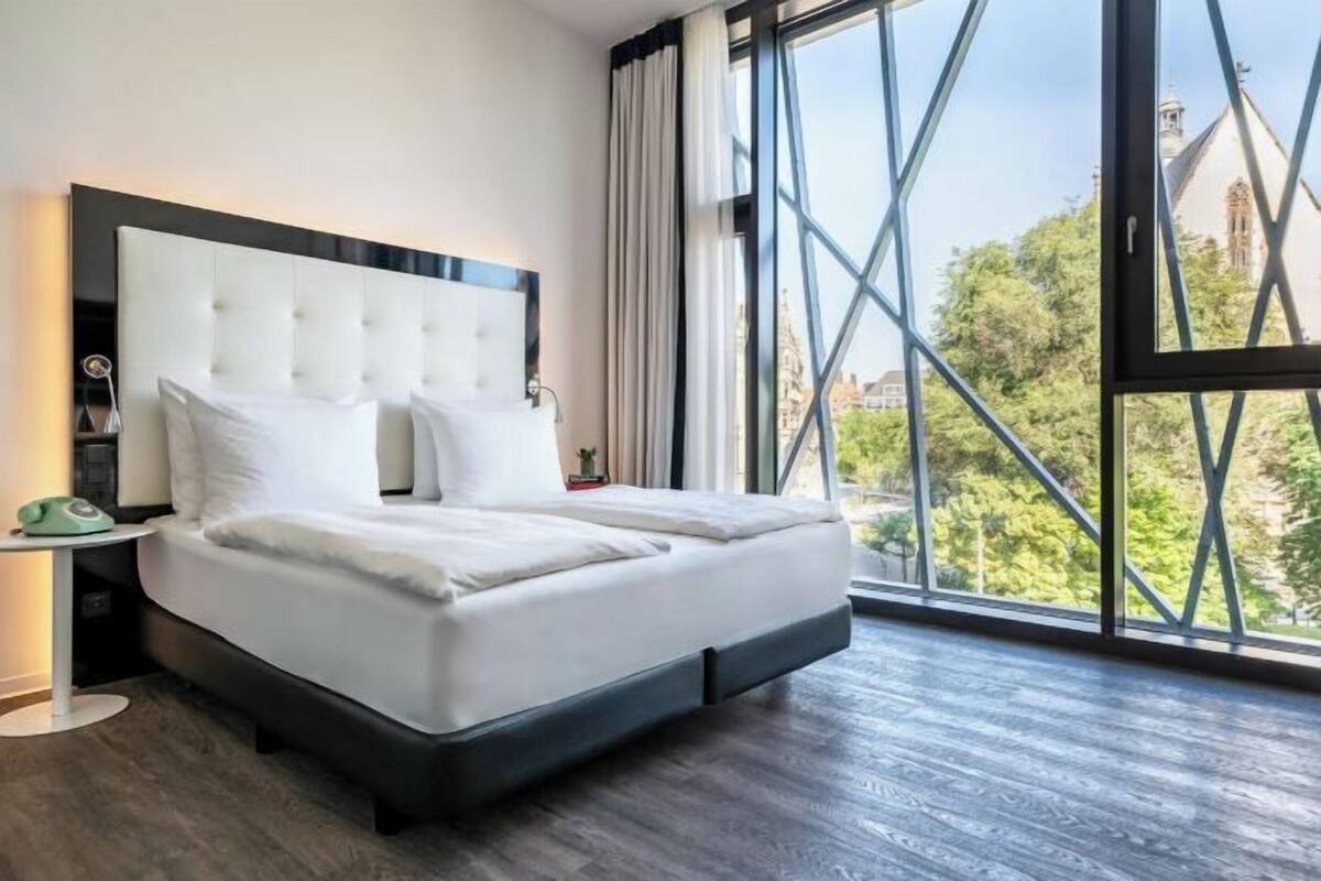 Een moderne hotelkamer met een groot bed, een nachtkastje, een lamp en een stadszicht door grote ramen.