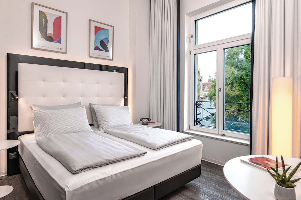 A modern hotel room with a large bed, a bedside table, a small table, a potted plant, and a window with a view of buildings.