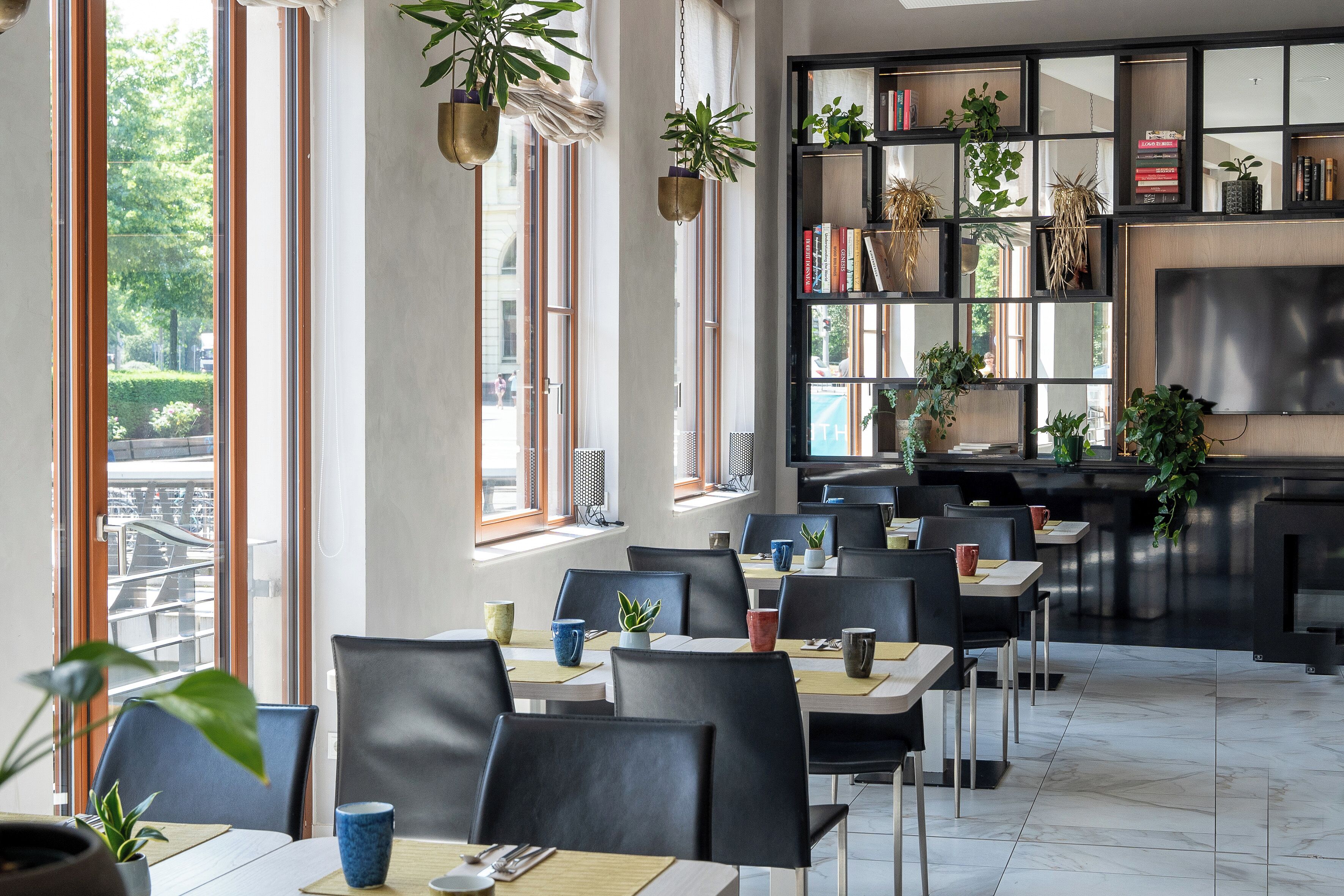 Ein modernes Restaurant mit mehreren Tischen und Stühlen, großen Fenstern und verschiedenen Kübelpflanzen.