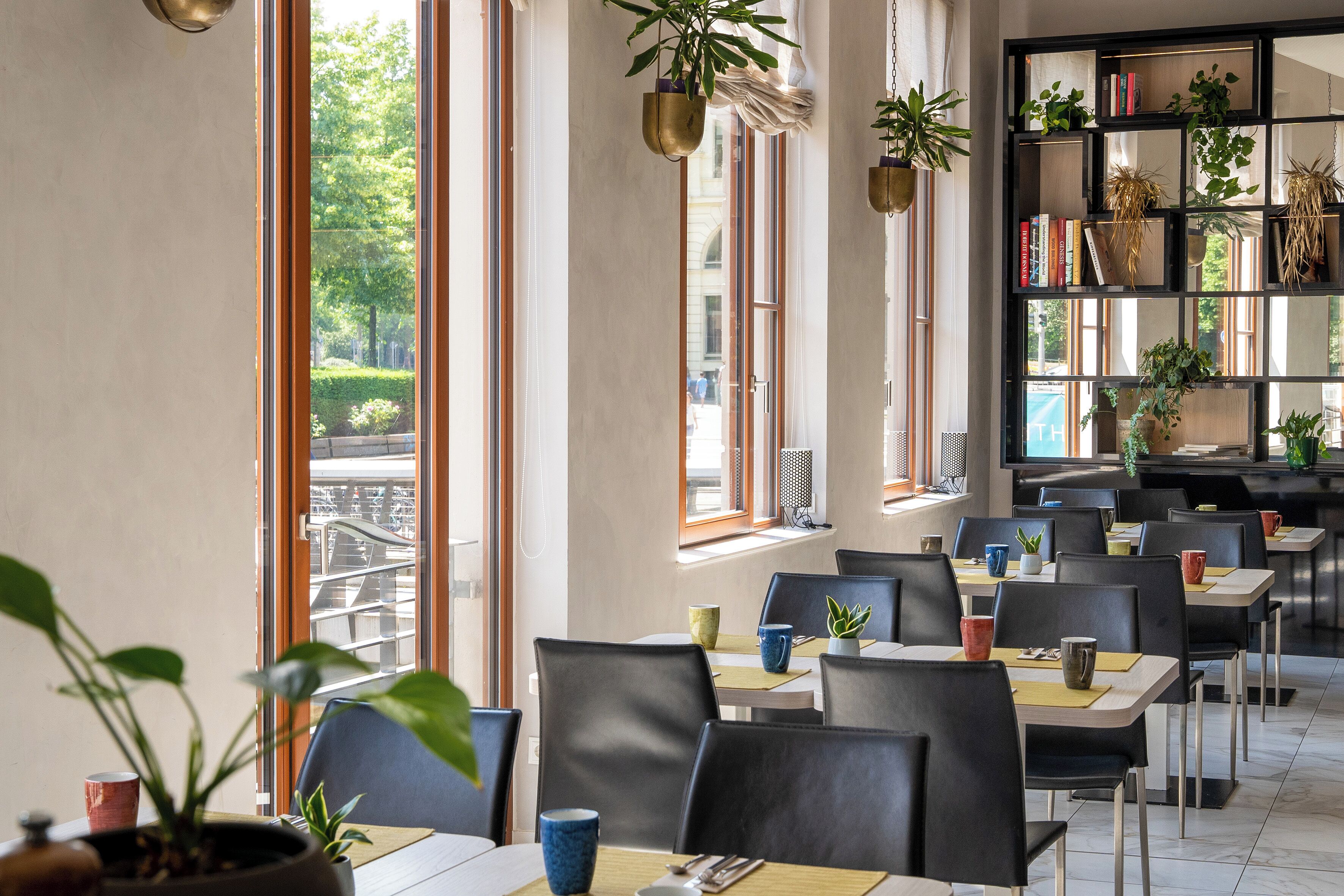 A restaurant with multiple tables and chairs, large windows with views of greenery, and shelves with plants and books.