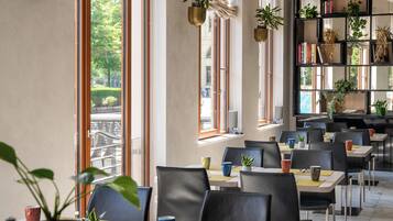 A restaurant with multiple tables and chairs, large windows with views of greenery, and shelves with plants and books.