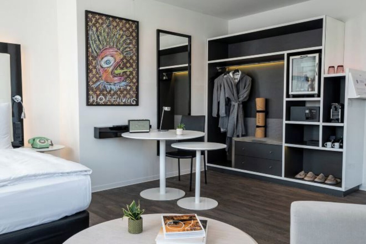 Ett modernt hotellrum med en säng, två vita, runda bord, en badrock som hänger på en klädställning och en stor spegel.