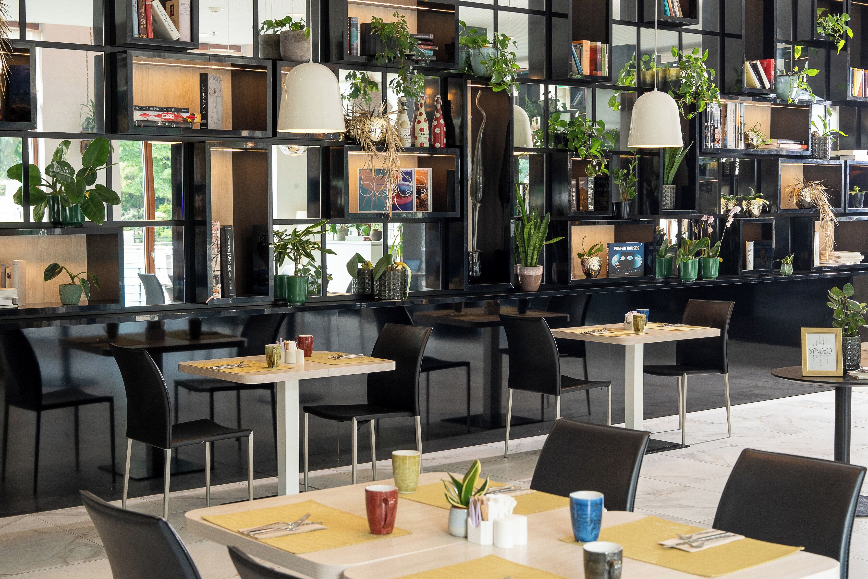 Ein modernes Restaurant mit schwarzen Regalen, die mit Büchern und Pflanzen gefüllt sind, Tischen mit Stühlen und einem klaren, zeitgemäßen Design.