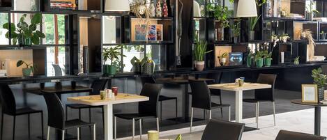 Ein modernes Restaurant mit schwarzen Regalen, die mit Büchern und Pflanzen gefüllt sind, Tischen mit Stühlen und einem klaren, zeitgemäßen Design.