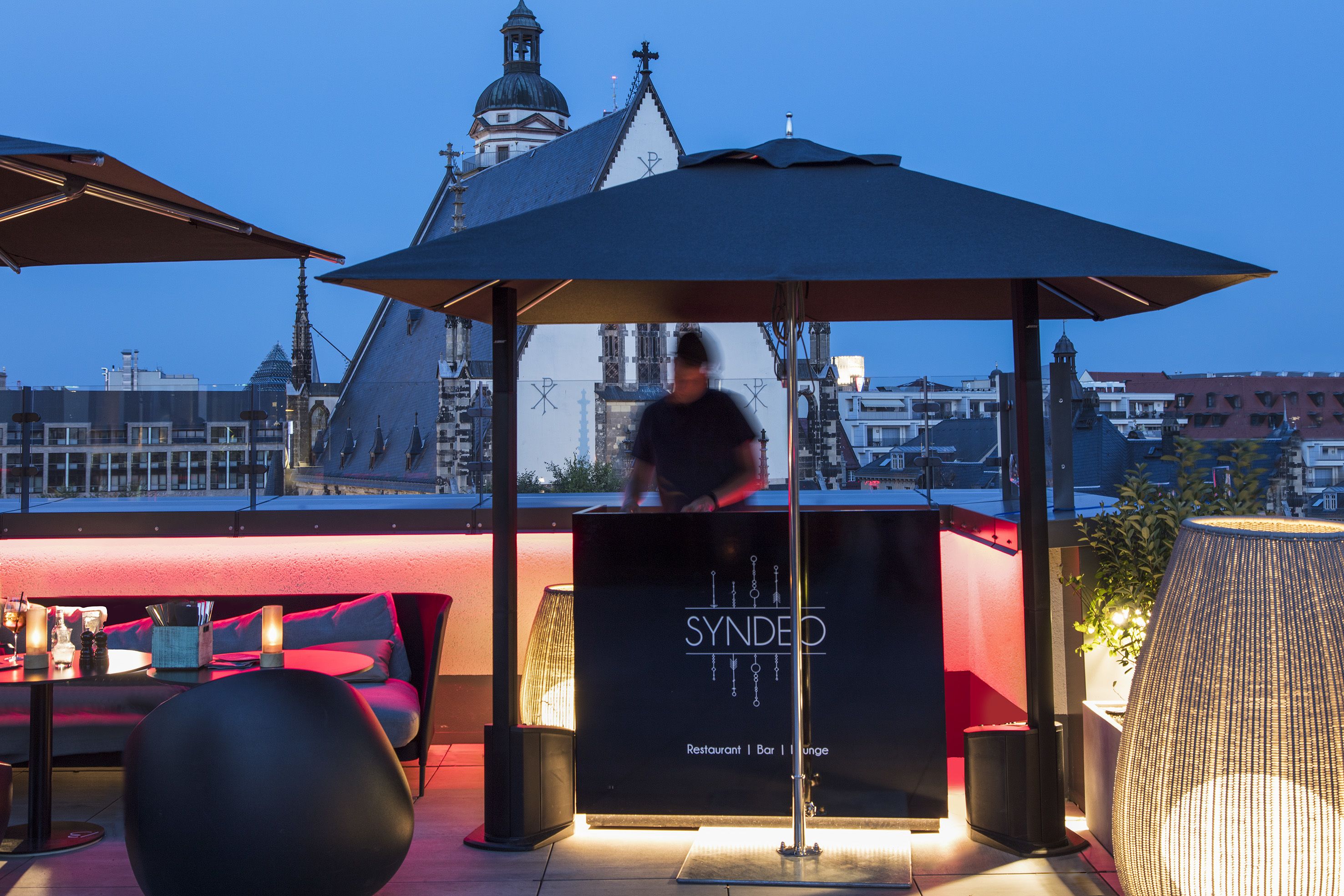 Eine SYNDEO Restaurant-Bar/Lounge mit modernem Design, mit einer Sitzecke mit runden Tischen und Stühlen und Blick auf die Stadtkulisse im Hintergrund.