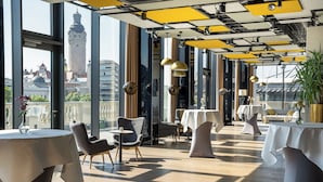 Un restaurante moderno con amplios ventanales, suelo de madera y vistas a una torre histórica. Las mesas están dispuestas con manteles blancos y sillas.