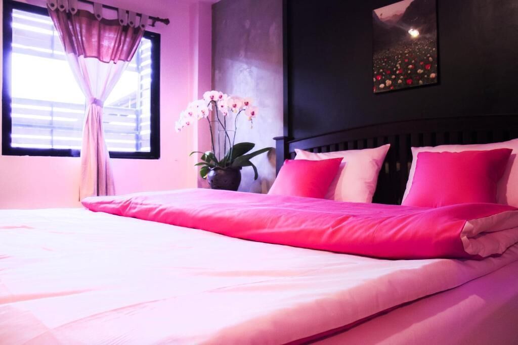 Un dormitorio con una cama grande, ropa de cama rosa, una ventana con cortinas, una planta en maceta y un cuadro colgado en la pared.