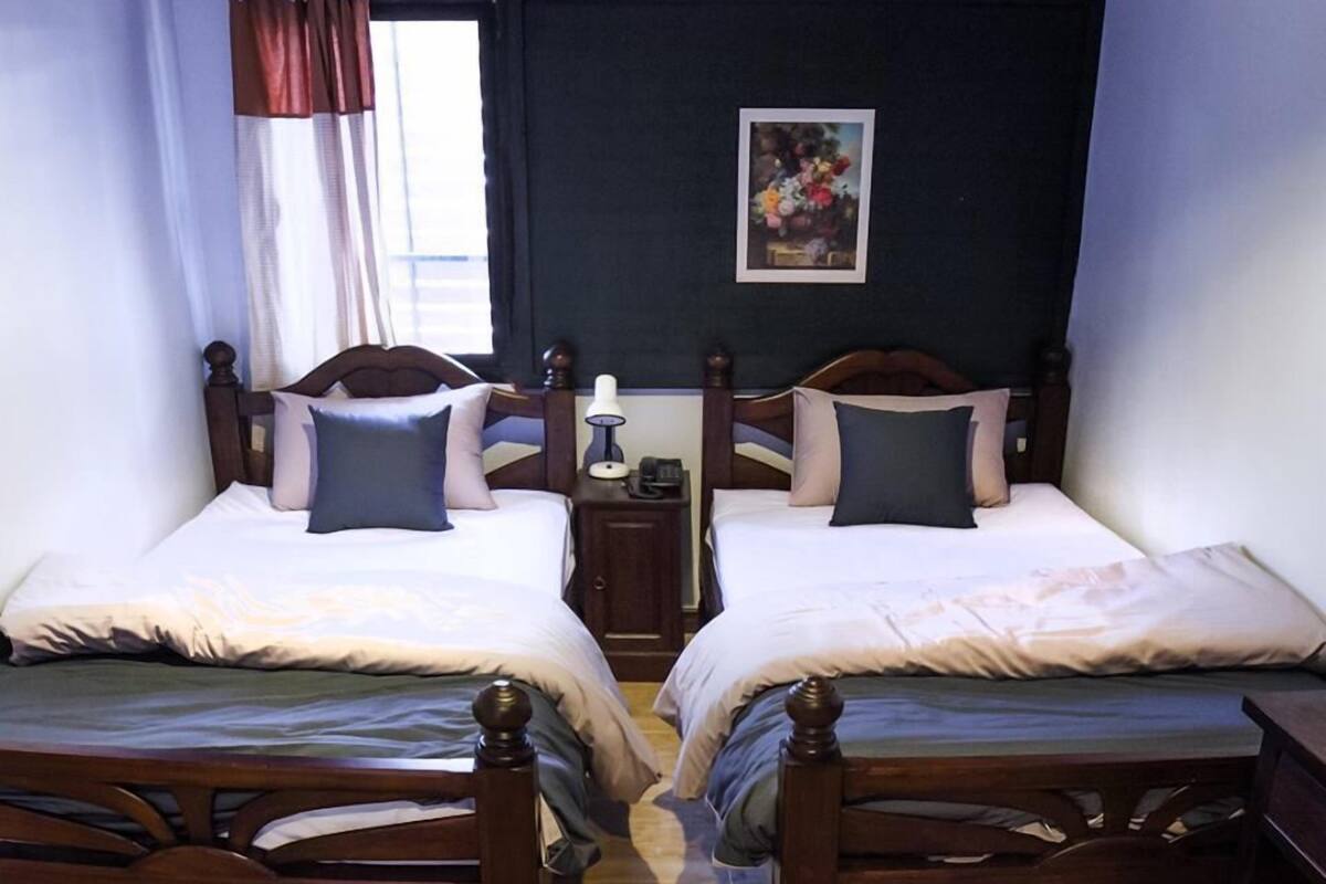 Quarto com duas camas de madeira, uma mesa de cabeceira com abajur, uma janela com cortinas e um quadro na parede.