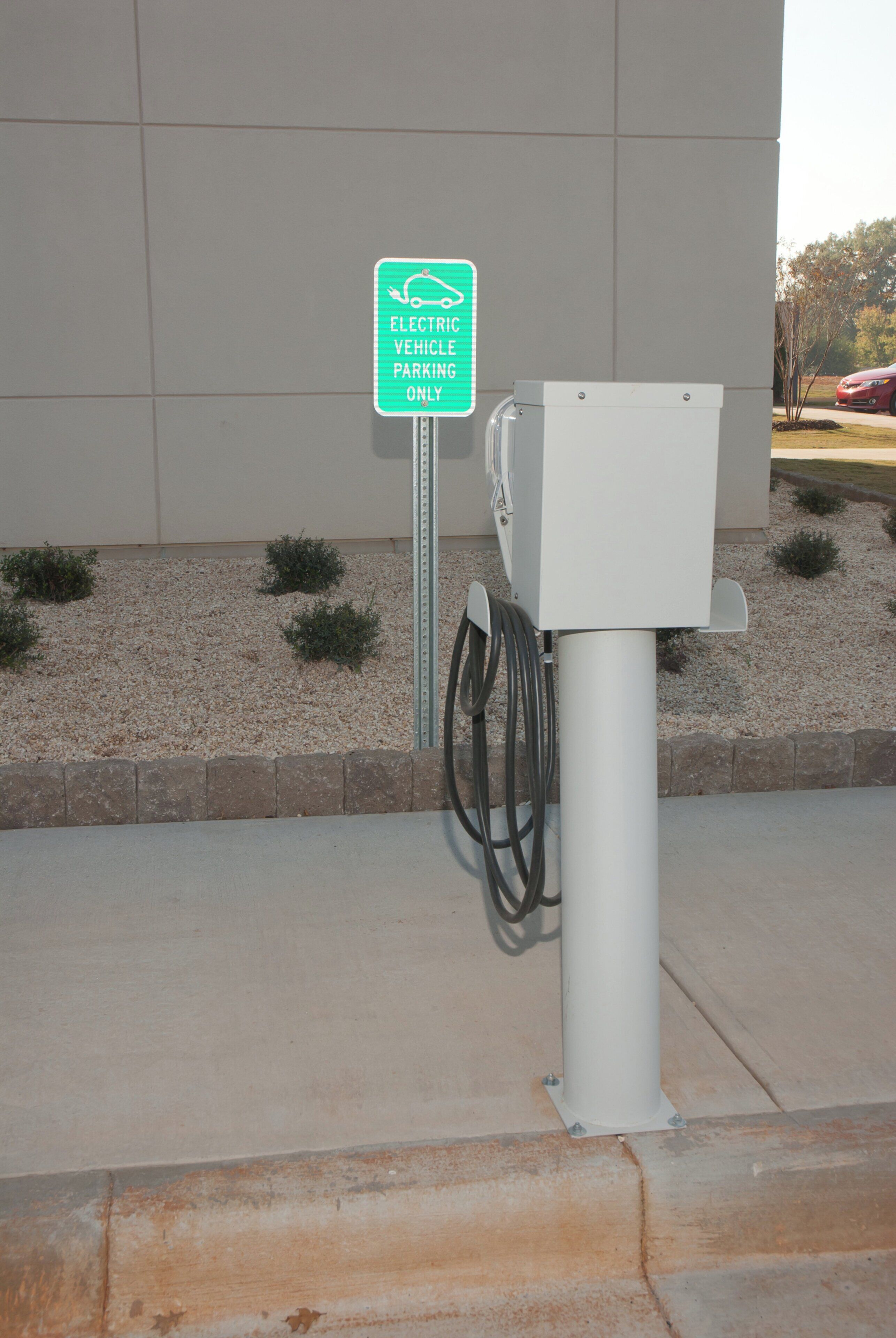 An electric vehicle charging station with a sign indicating it is for electric vehicles only, a cable connected to the charging unit, and a building in the background.