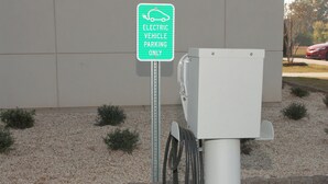 Une borne de recharge pour véhicules électriques, avec un panneau indiquant qu’elle est réservée aux véhicules électriques, un câble relié à l’unité de recharge et un bâtiment en arrière-plan.