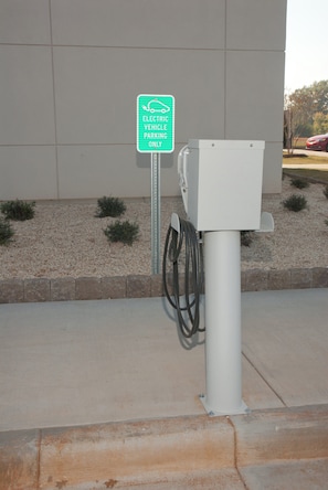 Une borne de recharge pour véhicules électriques, avec un panneau indiquant qu’elle est réservée aux véhicules électriques, un câble relié à l’unité de recharge et un bâtiment en arrière-plan.