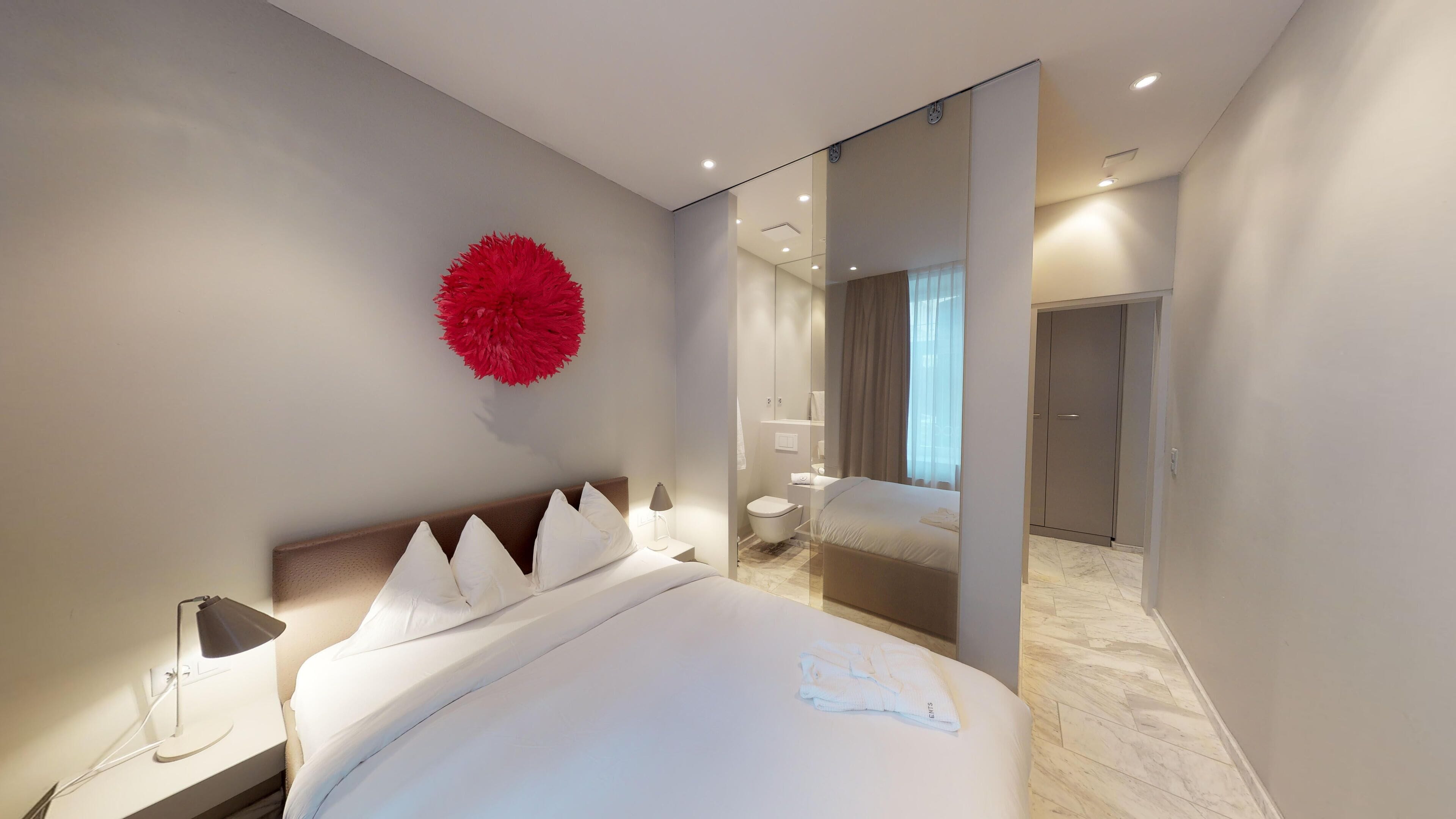 Ein modernes Hotelzimmer mit einem großen Bett, einem Nachttisch mit Lampe, einem Spiegel und einem roten dekorativen Element an der Wand.