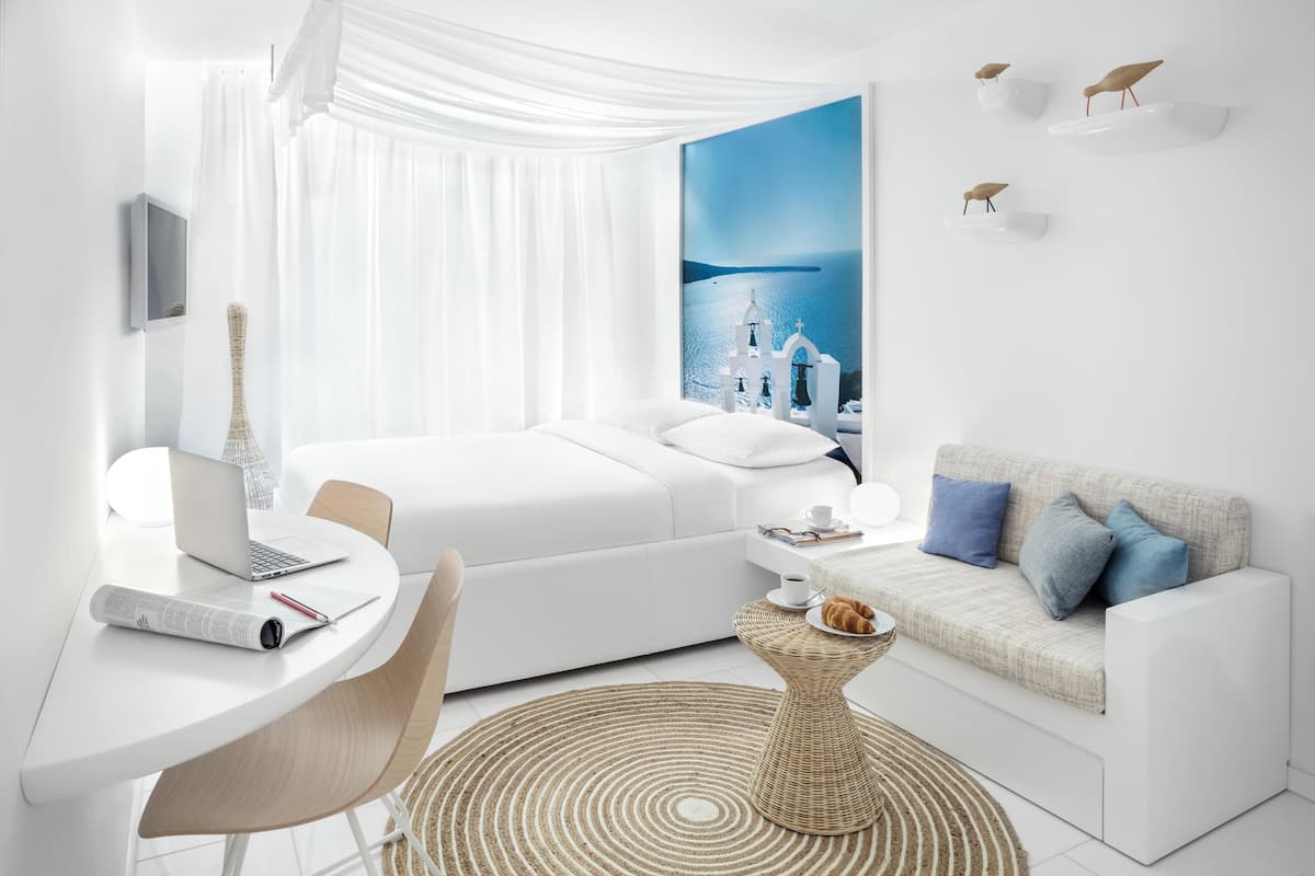 A modern hotel room with a bed, a desk with a laptop, a small round table with a croissant, and a view of a coastal scene with white buildings.