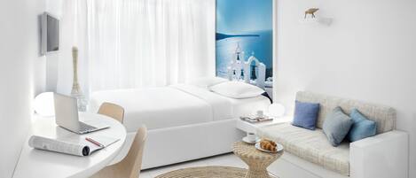 A modern hotel room with a bed, a desk with a laptop, a small round table with a croissant, and a view of a coastal scene with white buildings.