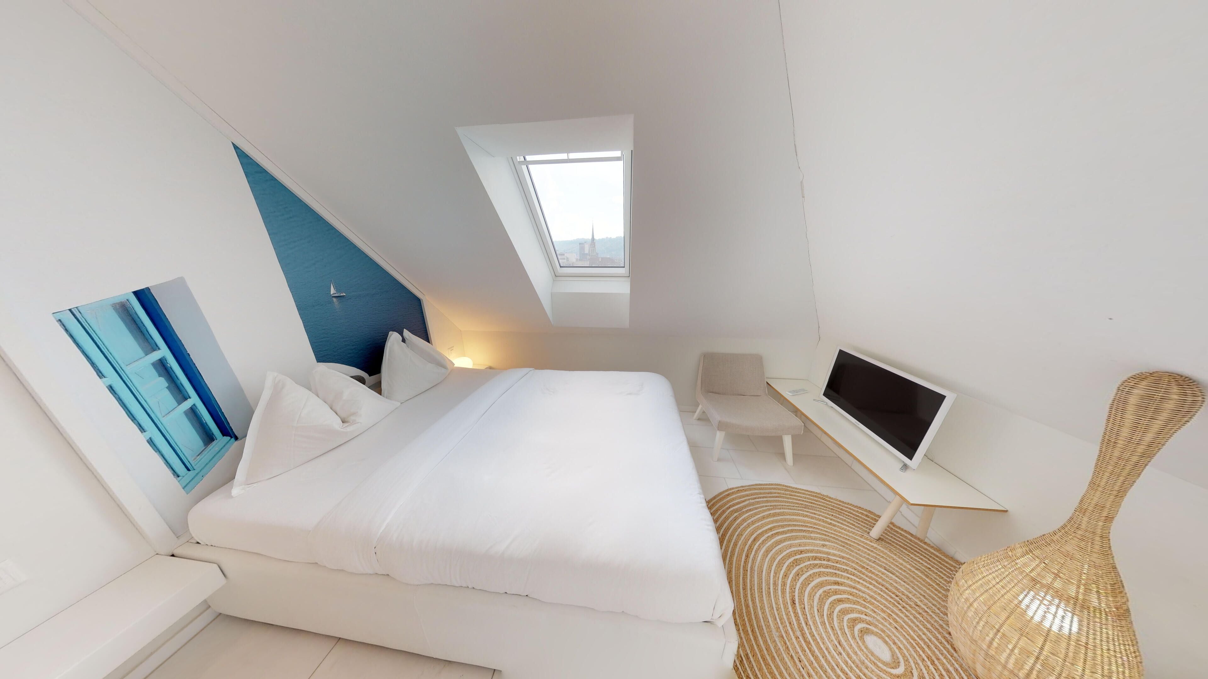 A modern bedroom with a large bed, a desk, a chair, a TV, and a view of the sea through a skylight.