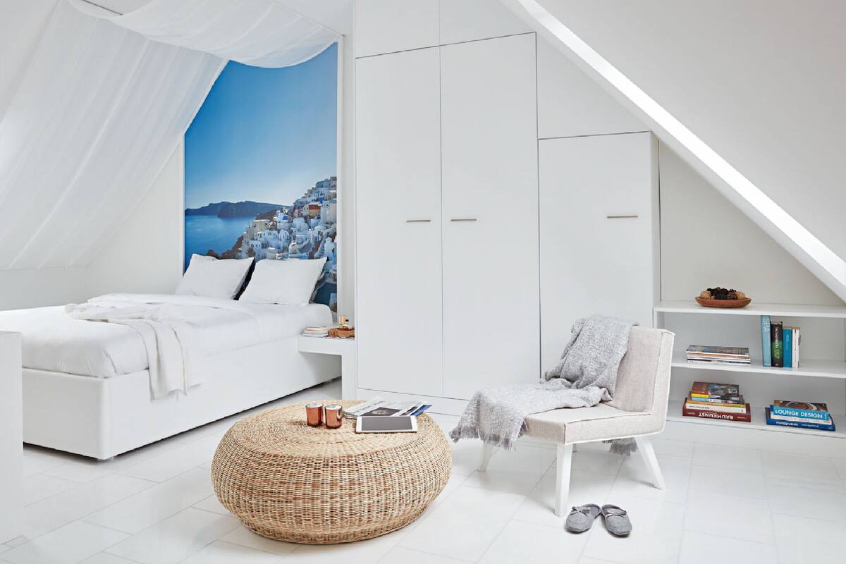 A modern bedroom with a white bed, a large window with a view of a coastal town, a white wardrobe, a reading chair, and a small bookshelf.