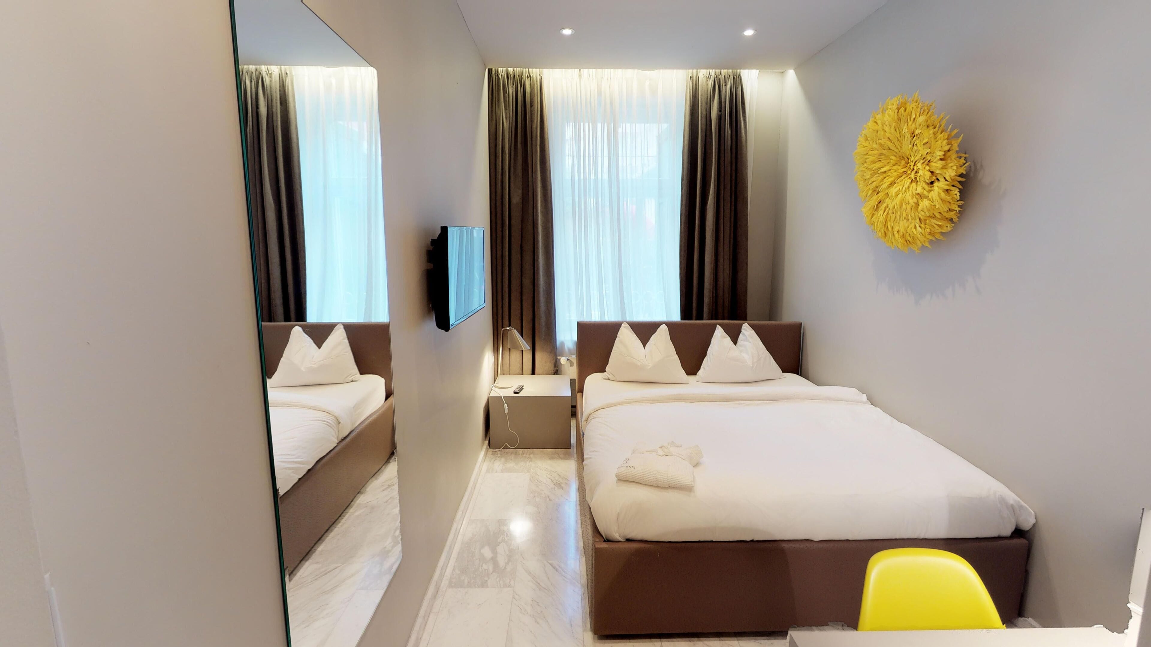 A modern hotel room with a large bed, a bedside table, a mirror, and a yellow chair.