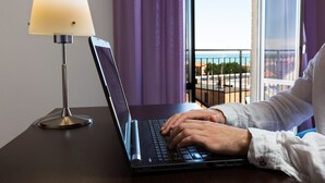 Un ordenador portátil sobre un escritorio con una lámpara, una ventana con cortinas y vistas a un balcón y edificios.