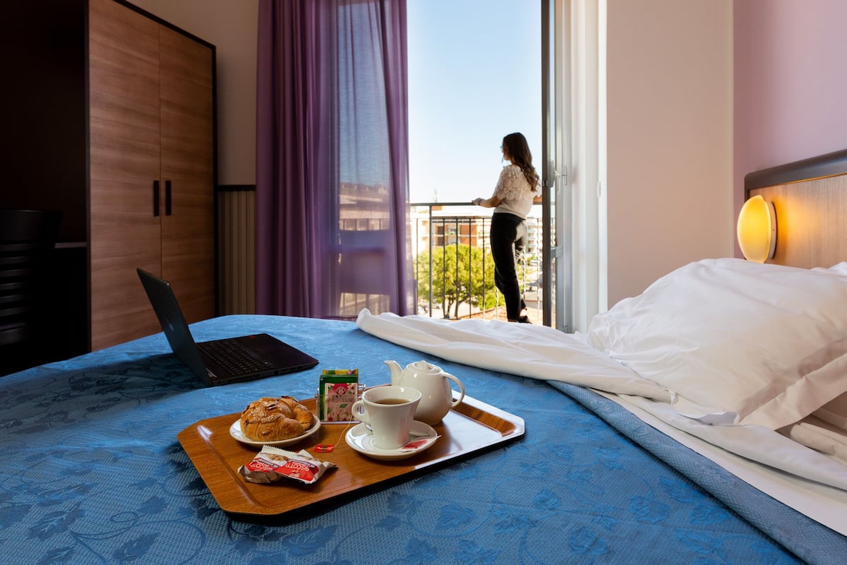 A hotel room with a bed, a laptop, breakfast tray, and a balcony with a person outside.