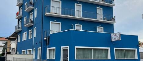 A blue multi-story hotel with balconies, a visible restaurant sign, and a pedestrian crossing in front.