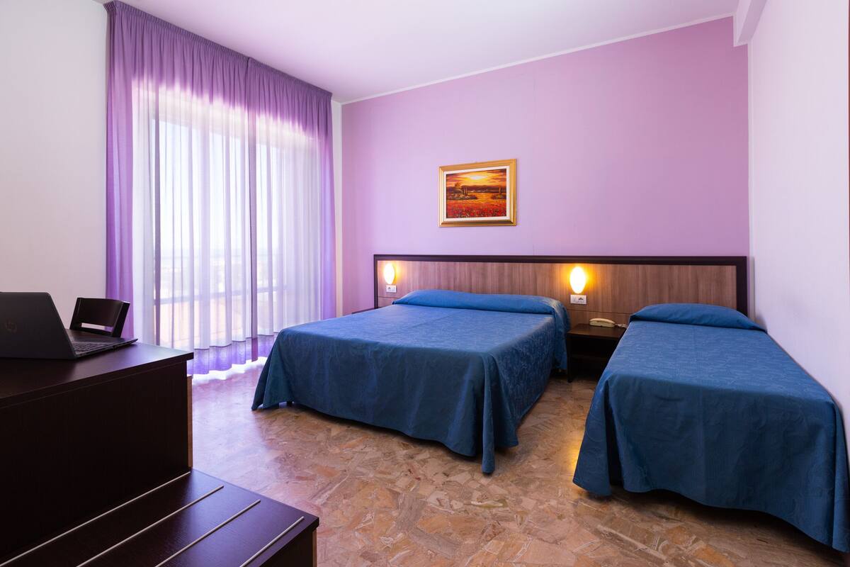 A hotel room with two beds, a desk with a laptop, a painting on the wall, and a window with purple curtains.