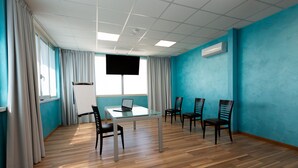 Un salón de conferencias con una mesa de vidrio, cuatro sillas, una pizarra, un televisor y una ventana con cortinas.