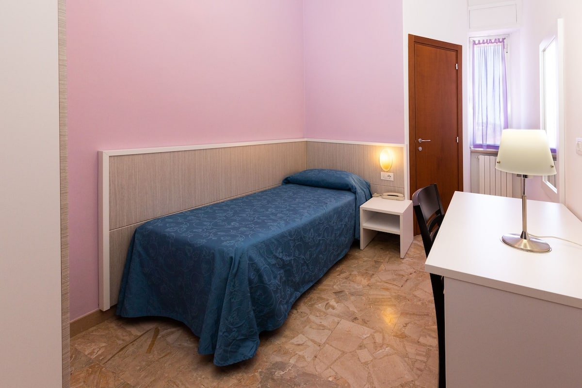 A small bedroom with a single bed, a nightstand, a desk, and a chair.