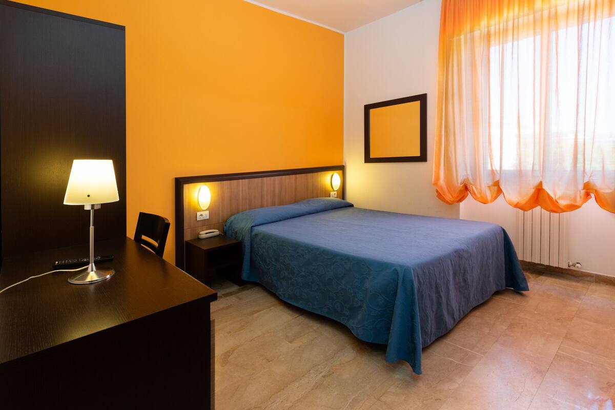 A hotel room with a bed, a desk with a lamp, a chair, and a window with curtains.
