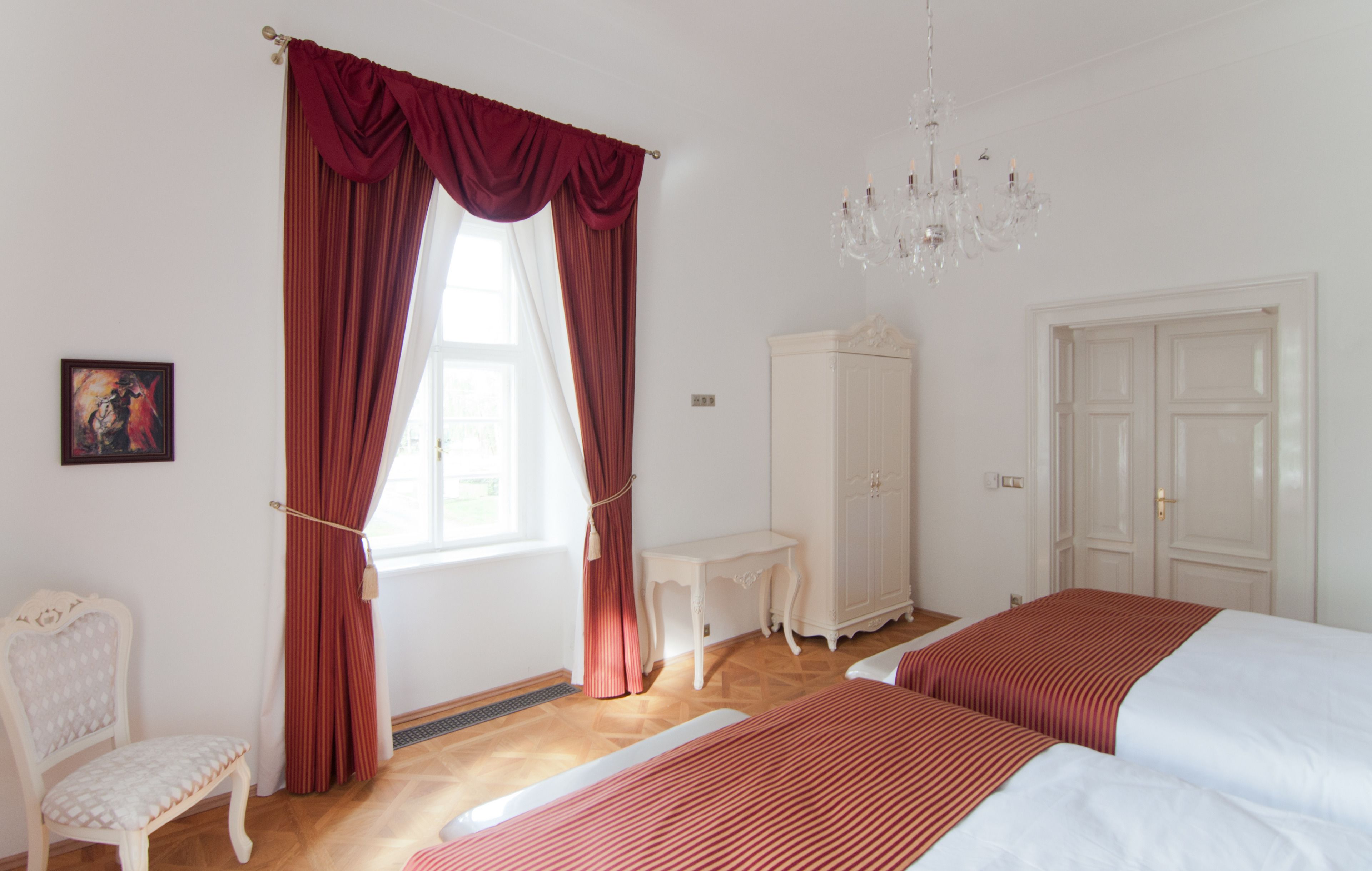 A bedroom with two beds, a white chair, a small table, a wardrobe, and a chandelier.