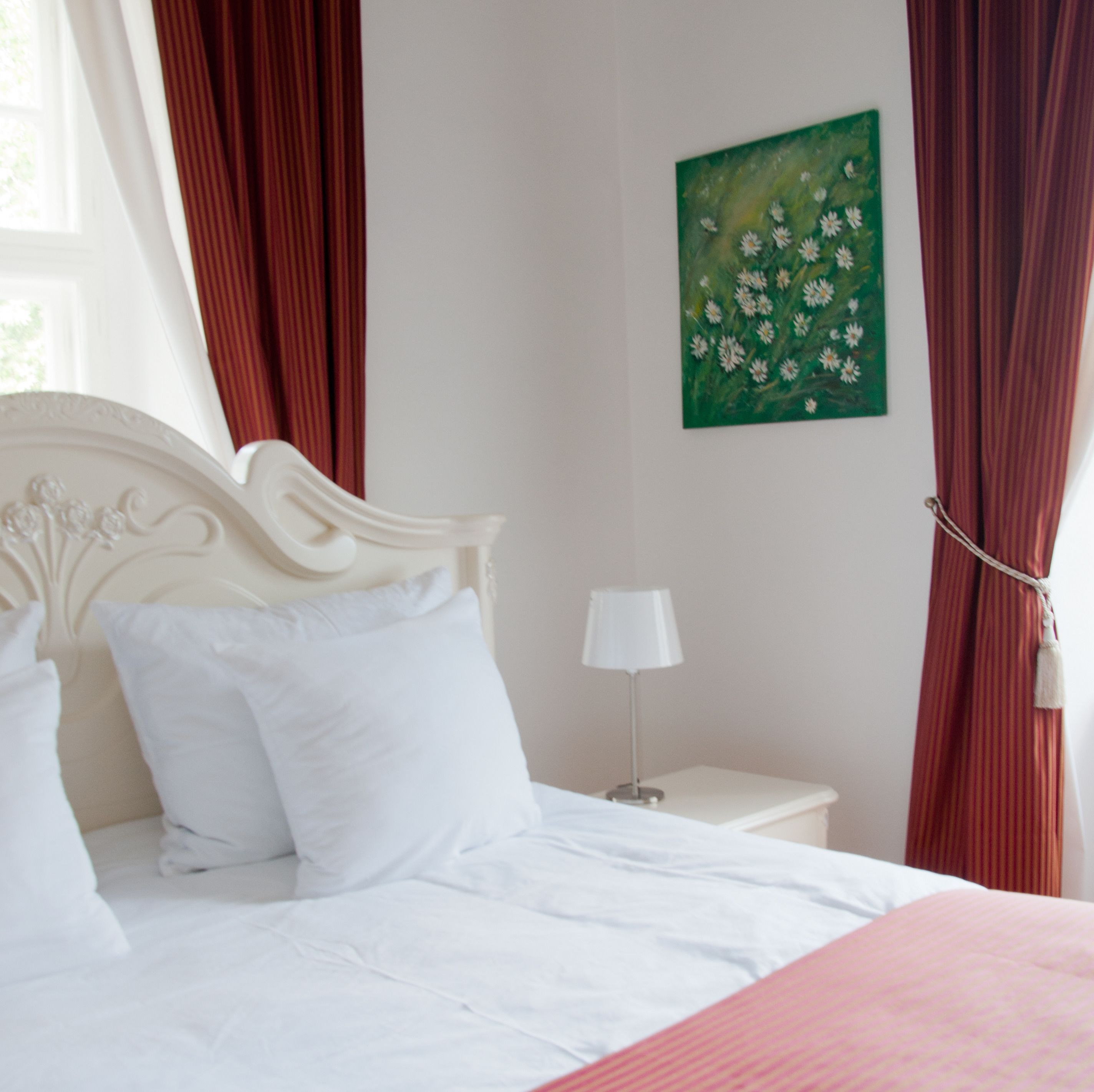 Ein ordentlich bezogenes Bett mit weißen Leinen, ein weißer Nachttisch mit Lampe, rote Vorhänge und ein Blumenbild mit Grün auf der Wand.