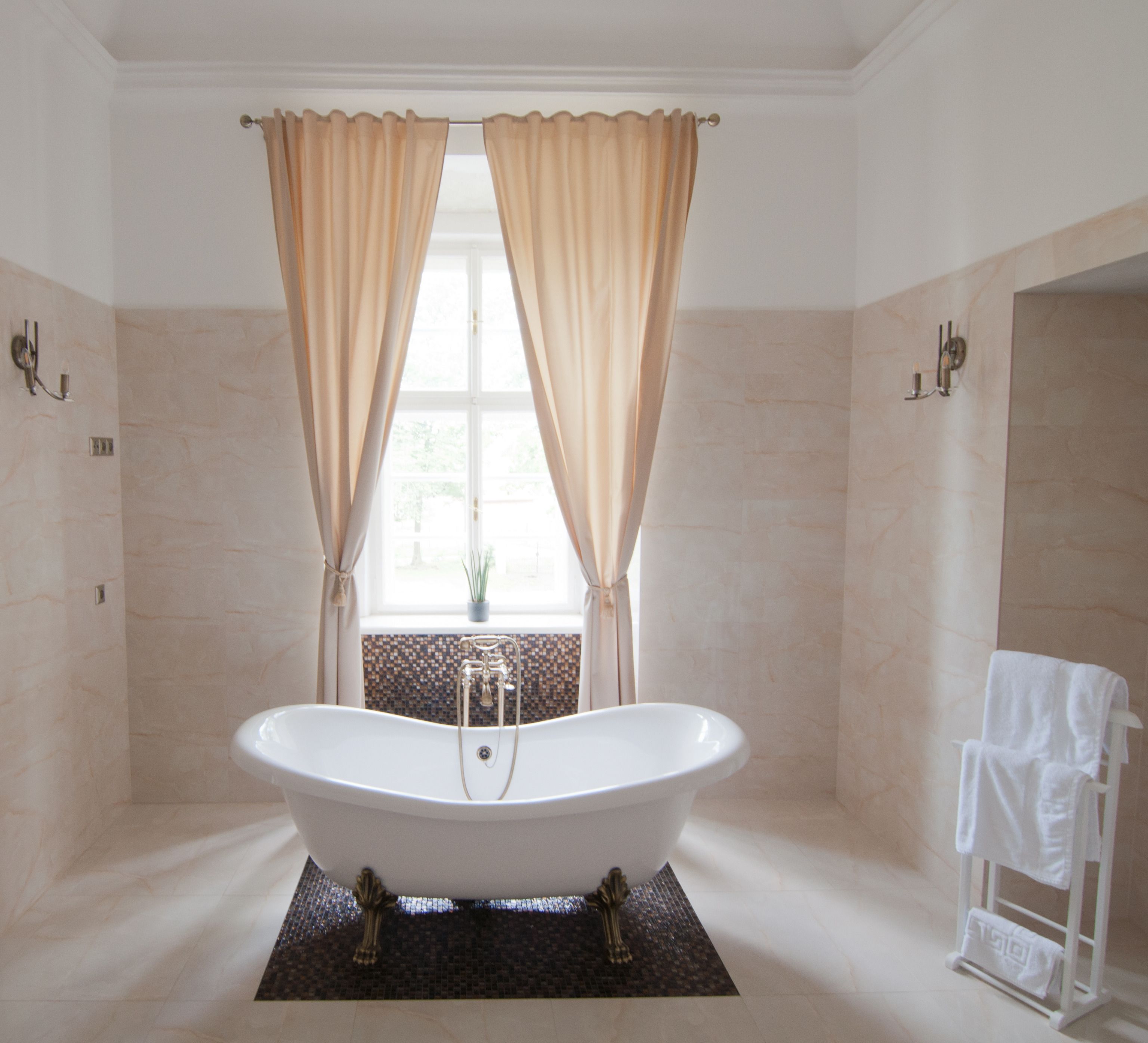 Un baño con una bañera independiente, cortinas color beige, un ventanal con vistas y un perchero para toallas.