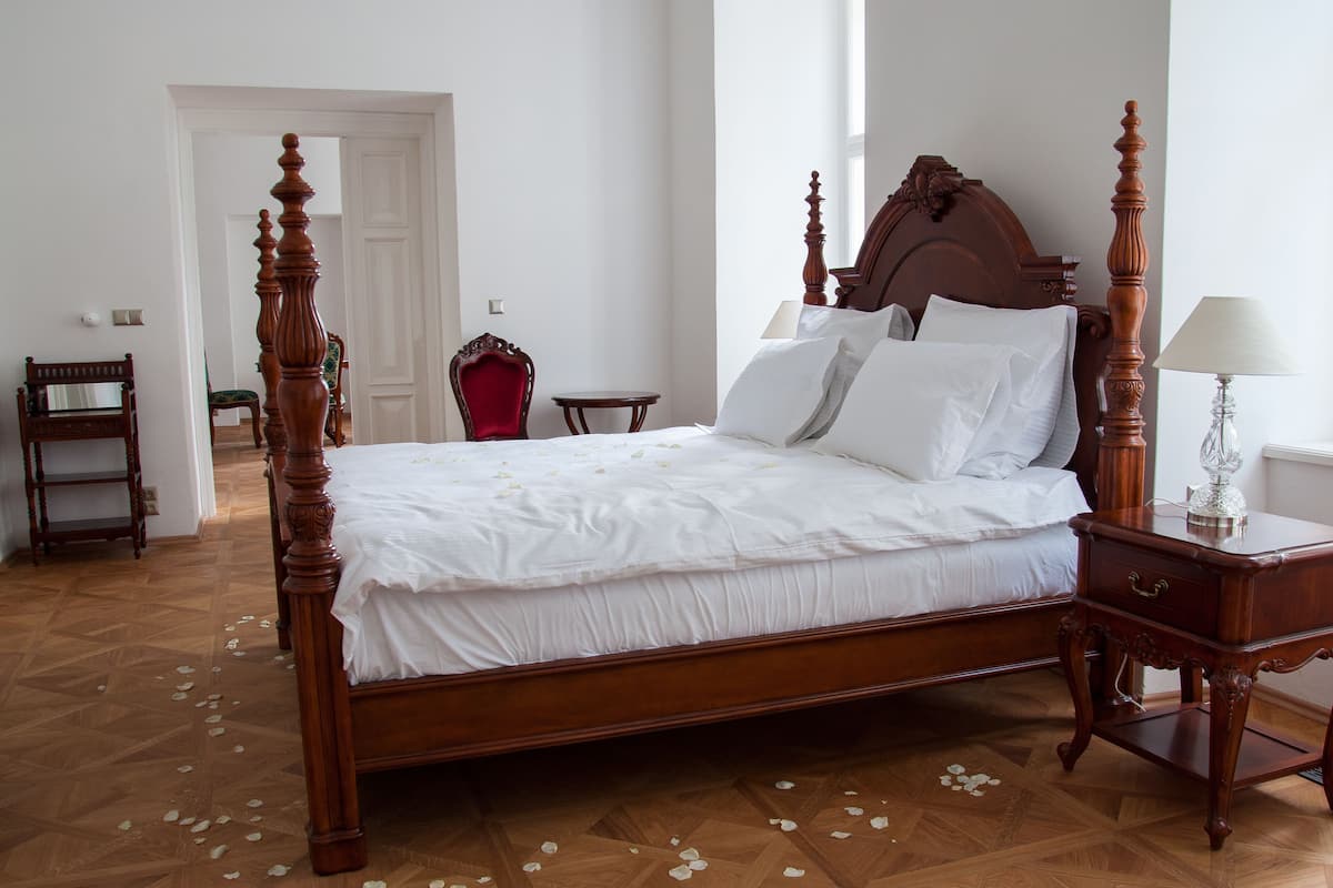 Ein großes hölzernes Bett mit weißen Leinen, ein Nachttisch aus Holz mit einer Lampe und ein roter Sessel in einem Raum mit Holzdielenboden und weißen Wänden.