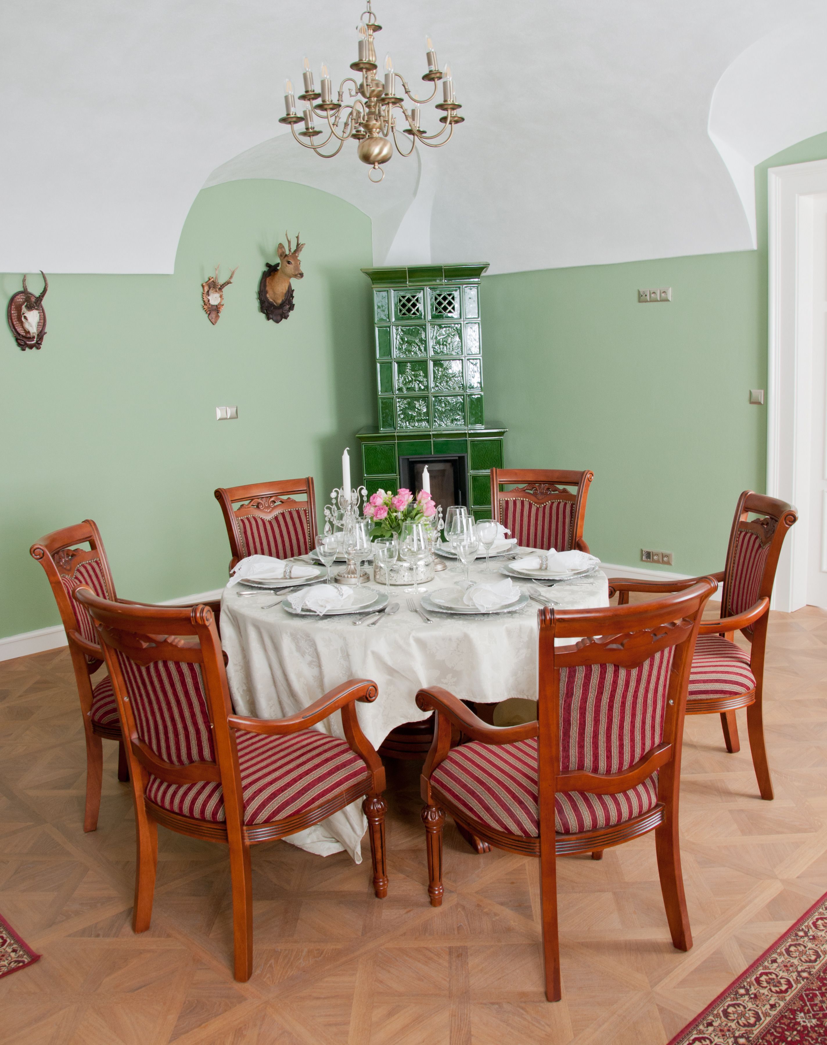 Ein Esszimmer mit einem runden Tisch, gedeckt für sechs Personen, umgeben von sechs Holzstühlen mit roten und weißen Streifen. Eine Kristallleuchter hängt von der Decke, und im Hintergrund ist ein grüner Kamin mit dekorativem Sims zu sehen.