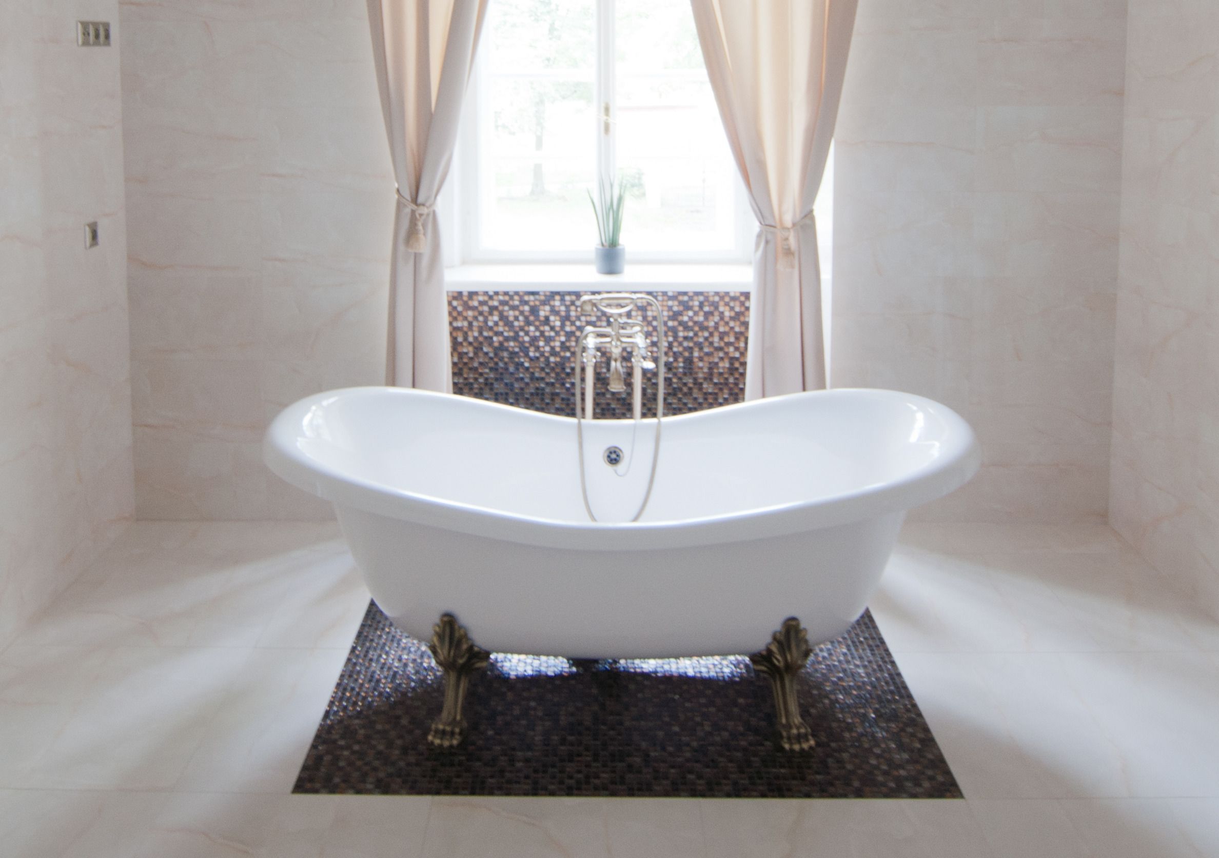 A freestanding bathtub in a bathroom with a tiled floor, a window with curtains, and a small plant on the windowsill.