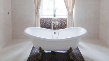 A freestanding bathtub in a bathroom with a tiled floor, a window with curtains, and a small plant on the windowsill.