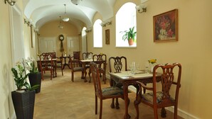 A dining area with wooden chairs and tables, a chandelier, and a large window with a plant. - Zámek Jeseník nad Odrou (Jesenik nad Odrou)