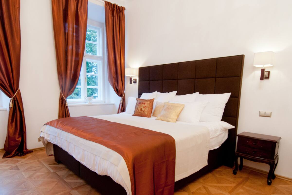 Ein ordentlich bezogenes Bett mit braunem Kopfteil, Holzboden und einem Fenster mit Vorhängen.