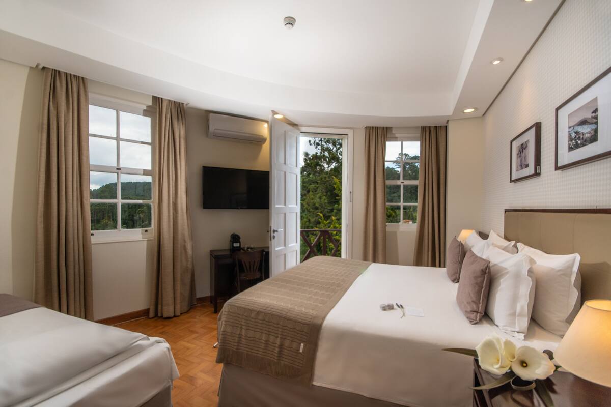 Habitación de hotel con cama, escritorio, silla, televisor y balcón con cortinas.