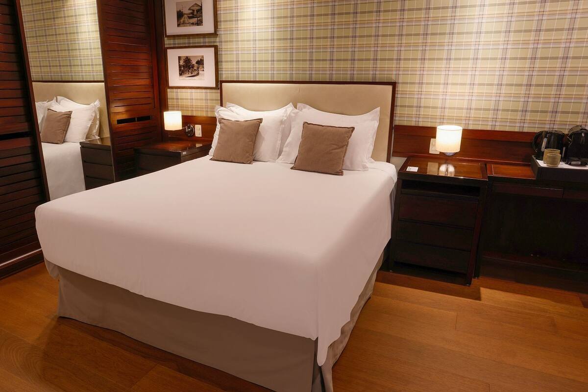 Habitación de hotel con una cama grande, cabecera de madera, mesita de noche con lámpara y espejo.