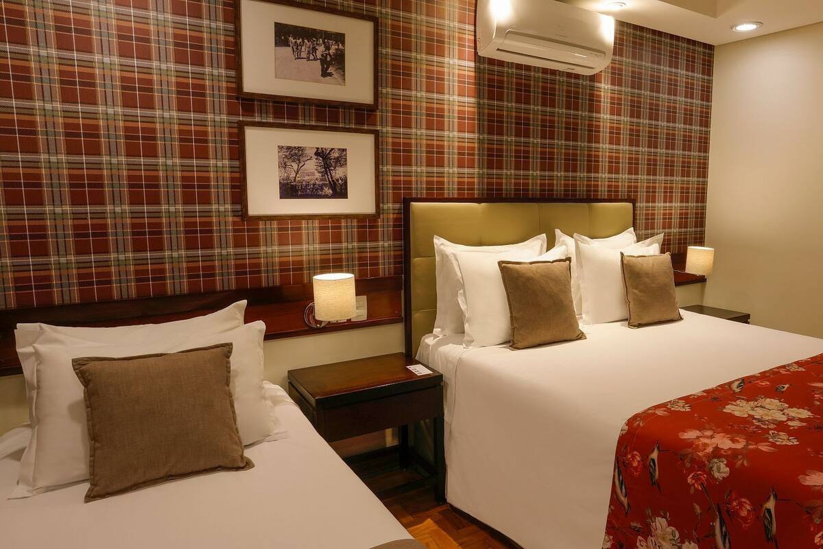 Habitación de hotel con dos camas, paredes de diseño escocés, cuadros enmarcados y una colcha de cama con estampado floral rojo.