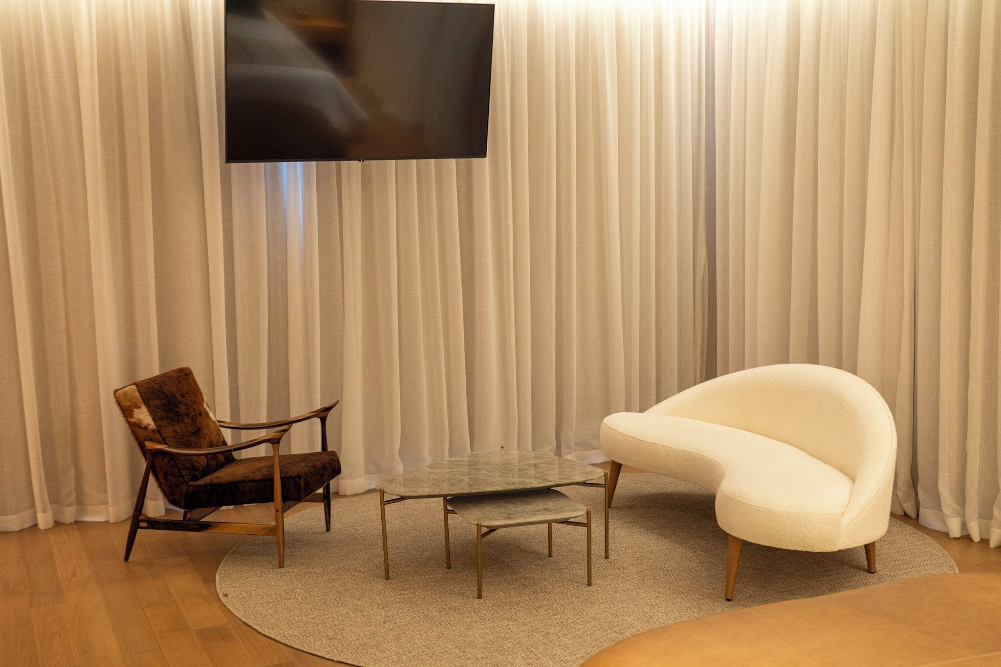 Una habitación con una televisión de pantalla plana montada en la pared, una mesa de centro redonda de cristal, un sillón blanco y una butaca de cuero marrón.