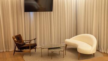 Una habitación con una televisión de pantalla plana montada en la pared, una mesa de centro redonda de cristal, un sillón blanco y una butaca de cuero marrón.