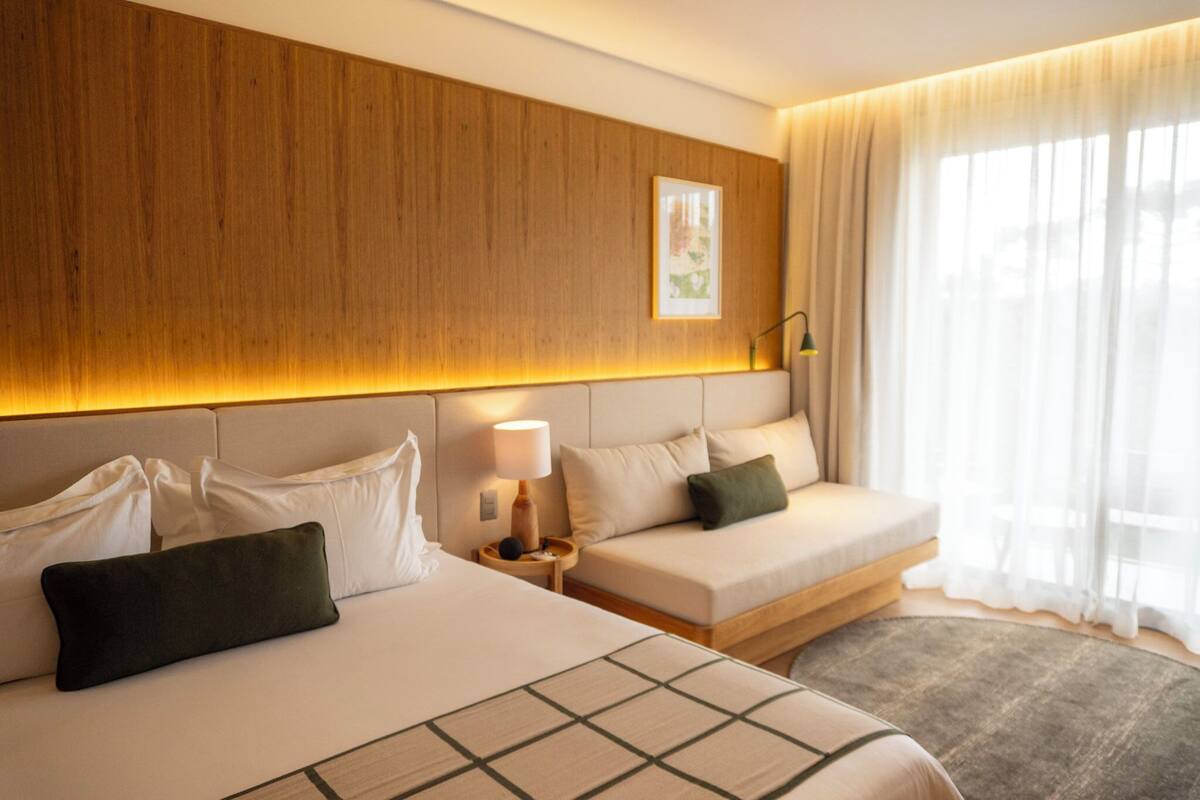 Habitación de hotel con cabecera de madera, cama con ropa de cama blanca, mesita de noche con lámpara y un ventanal con cortinas traslúcidas.