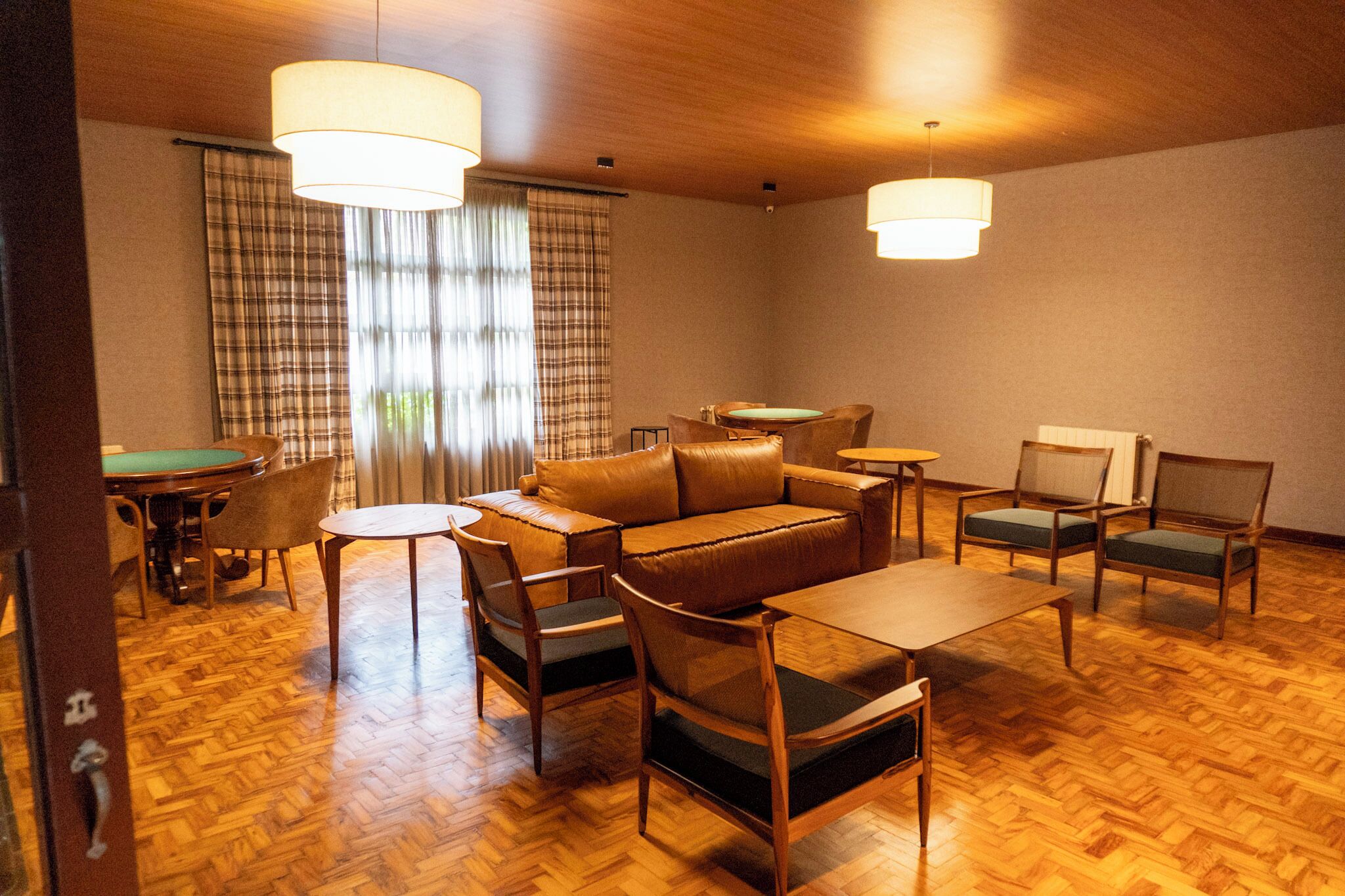 A room with a wooden herringbone floor, a brown leather sofa, two wooden tables, and four chairs with green cushions.