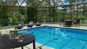 Área de piscina com paredes de vidro, cadeiras de descanso e uma mesa, com vista para uma floresta.